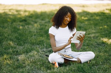 African girl holding a tablet for studying while sitting on a grass. Girl is using a white. Girl wearing a white clothes.