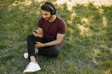 Young boy listening to music while sitting on a grass. Man using a smartphone. Man wearing a bordo shirt and jeans.