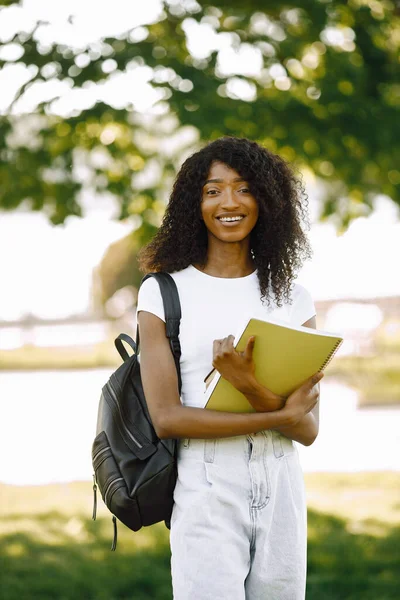African girl holding a books for studying while standing oudoors. Girl is looking directly at camera. Girl wearing a white clothes.