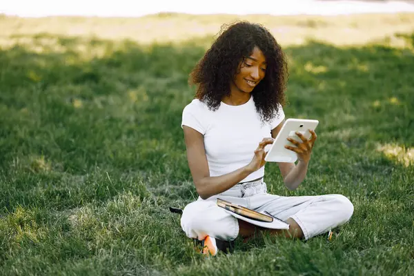 African girl holding a tablet for studying while sitting on a grass. Girl is using a white. Girl wearing a white clothes.