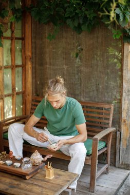 Blonde man sitting in outdoor Japanese spring garden in backyard porch of zen home. Man wearing green t-shirt and white trousers. Young man preparing for a tea ceremony.