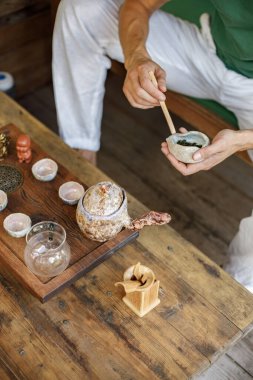 Cropped photo of a man preparing for a tea ceremony. Man sitting in outdoor Japanese spring garden in backyard porch of zen home. Focus on a cup of tea.