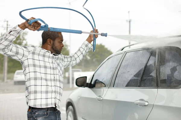 Man washing luxury car on a car wash with a pressure washer. Black man using high pressure hose with water. Man wearing plaid shirt.
