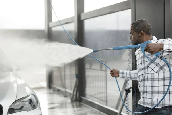 Man washing luxury car on a car wash with a pressure washer. Black man using high pressure hose with water. Man wearing plaid shirt.