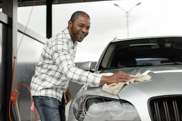 Man washing luxury car on a car wash with a rag. Black man wiping his car. Man wearing plaid shirt, smiling and looking at camera.