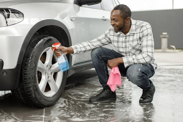 Man washing luxury car on a car wash using sprayer bottle and a rose rag. Black man polishing his car. Man wearing plaid shirt and jeans.