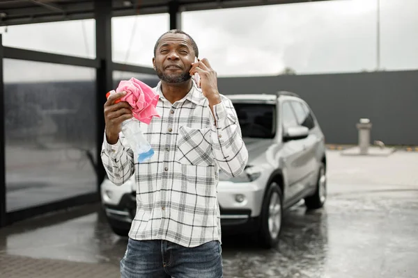 Man washing luxury car on a car wash using sprayer bottle and a rag. Black man talking on the phone and holding in his hands rose rag and sprayer bottle for polishing his car. Man wearing plaid shirt