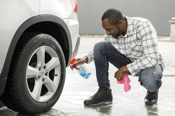 Man washing luxury car on a car wash using sprayer bottle and a rose rag. Black man polishing his car. Man wearing plaid shirt and jeans.