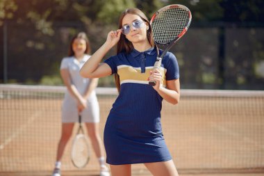 Two girls playing on a tennis court. Women wearing white and blue sport dresses. Focus on a girl in blue dress and sunglasses, who looking at camera.