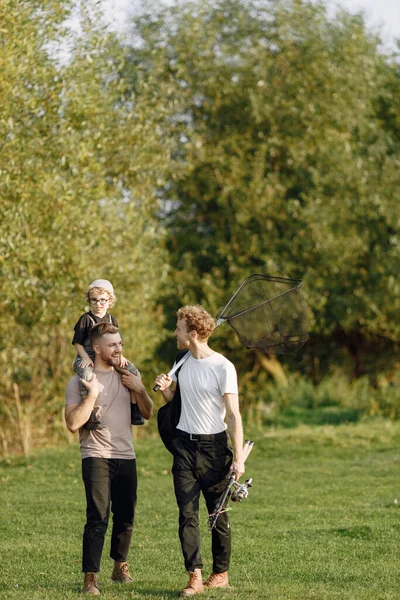 stock image Dad, his friend and his child boy spend time outdoors together. Curly toddler boy wearing a khaki overall. Boy sitting on a shoulders of his father.