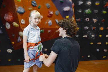 Eğitmen, küçük bir kızın içerideki kaya tırmanış merkezinde emniyet kemeri takmasına yardım ediyor. Küçük kız, çocuk eğlence merkezinde kaya tırmanışı yapıyor. Sarışın Kafkasyalı