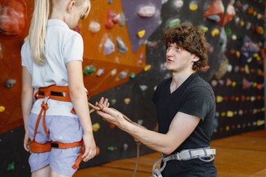 Eğitmen, küçük bir kızın içerideki kaya tırmanış merkezinde emniyet kemeri takmasına yardım ediyor. Küçük kız, çocuk eğlence merkezinde kaya tırmanışı yapıyor. Sarışın Kafkasyalı