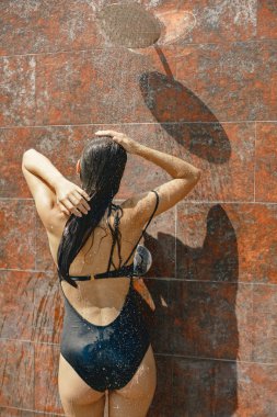 Mayo giymiş esmer bir kadının arka görüntüsü açık havada duş alıyor. Siyah mayo giyen bir kız. Fotoğraf için poz veren uzun saçlı genç bir kadın..