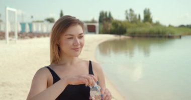 Siyah elbiseli genç kadın nehir veya gölün arka planındaki şeffaf şişeden su içiyor. Çekici kız açık hava sporlarından sonra sıvı içiyor..