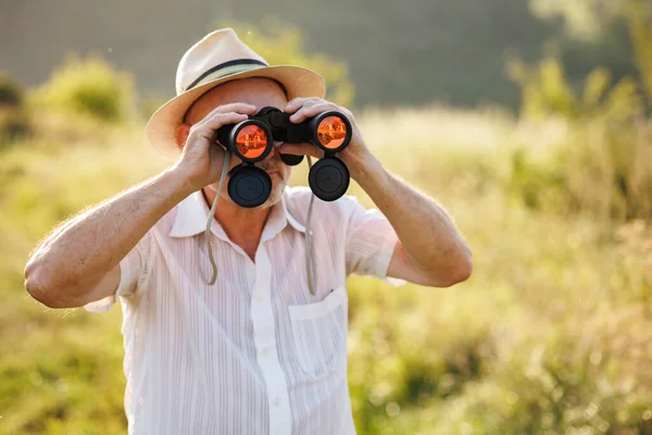Portrait of a mature man with binoculars in a field. Bearded caucasian man looking through a binoculars in a field at summer. Man wearing striped t-shirt and a hat.