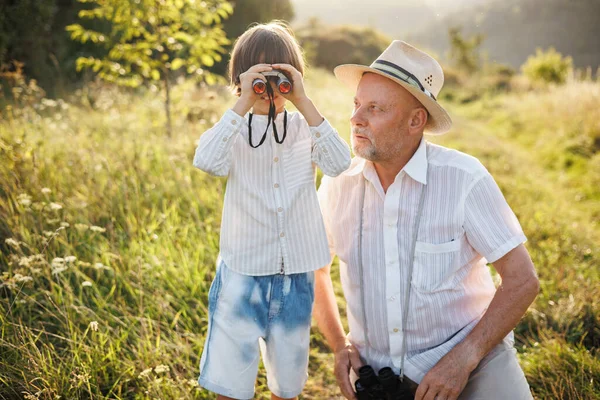 stock image Portrait of little explorer and his grandfather with binoculars in a field. Boy and grandfather spending time together at summer. Brunette boy and man wearing striped shirts.