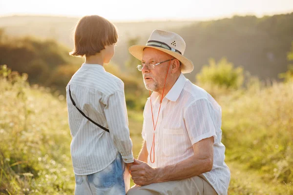 Photo of little boy and his grandfather standing and talking in the field. Boy and grandfather spending time together at summer. Brunette boy and man wearing striped shirts.