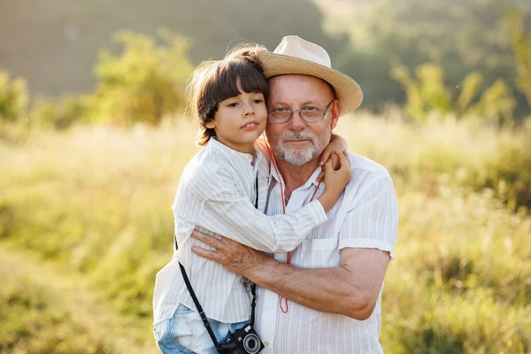 Photo of little boy and his grandfather standing and hugging in the field. Boy and grandfather spending time together at summer. Brunette boy and man wearing striped shirts.