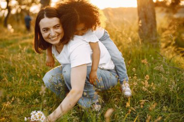 Kafkasyalı annenin Afrikalı Amerikalı kızını sırtında taşıdığı fotoğraf. Kızın siyah kıvırcık saçları var. Anne ve kızı beyaz tişört giyiyor..