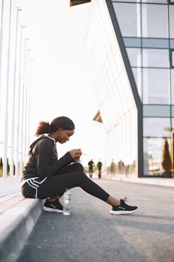 Afro-amerikan fitness modeli açık hava eğitimi. Siyah spor giyimli bir kadın. Kaldırımda oturan ve telefona bakan bir kadın..