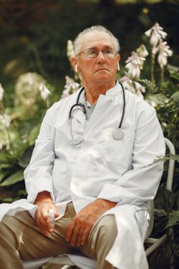 Doctor in a white uniform. Old man sitting in a summer park. Senior with stethoscope.