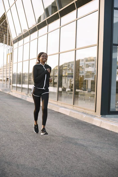African american fitness model jogging outdoors. Woman wearing black sportswear. Woman running near glass building.