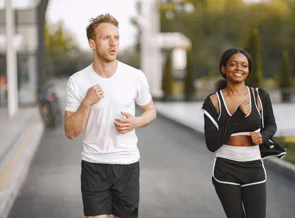 African american fitness model and caucasian man jogging outdoors. Woman wearing black sportswear. Man wearing white t-shirt.