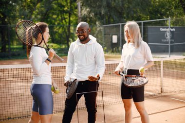 Kız arkadaşlar tenis dersi alıyor. Siyahi adam koçu konuşuyor ve nasıl oynanacağını anlatıyor. Açık hava çocuk parkı.