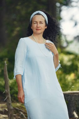 Kıdemli kadın kameraya bakıyor. Mavi elbiseli kadın. Esmer yürür.