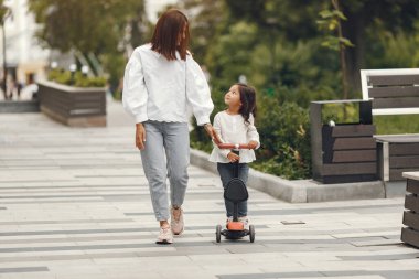 Anne ve kızı parkta scooter 'a biniyor. Çocuklar paten kaymayı öğreniyor. Küçük kız güneşli yaz gününde kayıyor.