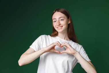 Yeşil arka planda izole edilmiş beyaz bir kızın kapalı bir portresi. Beyaz tişörtlü, fotoğraf için poz veren ve elleriyle kalbi gösteren güzel bir kadının portresi. Genç bir kızın stüdyo fotoğrafı..