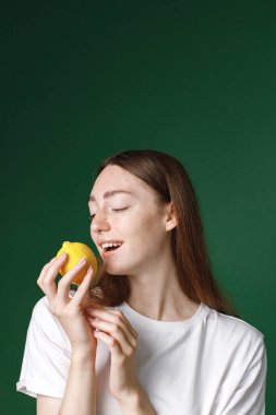 Yeşil arka planda izole edilmiş beyaz bir kızın kapalı bir portresi. Beyaz tişörtlü güzel bir kadının portresi neşe içinde fotoğraf için poz veriyor. Limon tutan genç bir kızın stüdyo fotoğrafı..