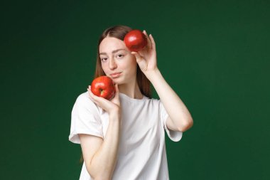 Yeşil arka planda izole edilmiş beyaz bir kızın kapalı bir portresi. Beyaz tişörtlü güzel bir kadının portresi neşe içinde fotoğraf için poz veriyor. İki kırmızı elma tutan genç bir kızın stüdyo fotoğrafı..