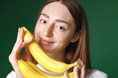 Yeşil arka planda izole edilmiş beyaz bir kızın kapalı bir portresi. Beyaz tişörtlü güzel bir kadının portresi neşe içinde fotoğraf için poz veriyor. Üç muz tutan genç bir kızın stüdyo fotoğrafı..