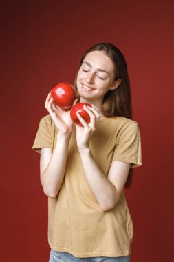 Kırmızı arka planda izole edilmiş beyaz bir kızın stüdyo portresi. Bej tişörtlü, fotoğraf için poz veren güzel bir kadının portresi. İki kırmızı elma tutan genç bir kızın stüdyo fotoğrafı..