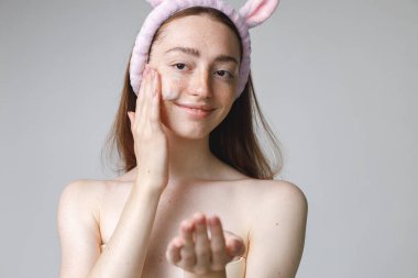 Çekici bir genç kadının güzel portresi beyaz arka planda yüzüne krem sür. Kadının gömleği yok. Kadının uzun kızıl saçları ve tavşan kulaklı saç bandı var..