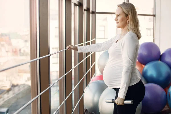 Caucasian pregnant woman in third trimester doing fitness excercise indoors. Woman in bright fitness studio with big windows. Blonde woman wearing whit shirt and black leggins.