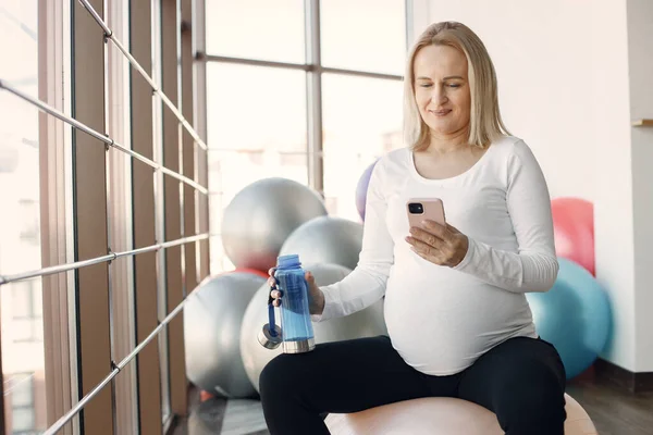 Caucasian pregnant woman in third trimester using a phone a holding a bottle with water. Woman in bright fitness studio with big windows. Blonde woman wearing whit shirt and black leggins.