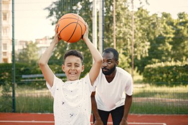 Genç babası ve oğlu parkın yakınındaki basketbol sahasında basketbol oynuyorlar. Beyaz tişört giyen adam ve çocuk. Çocuğun gözleri kapalı..