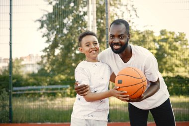 Genç babası ve oğlu parkın yakınındaki basketbol sahasında basketbol oynuyorlar. Beyaz tişört giyen adam ve çocuk. Çocuklar topu tutuyor ve kameraya bakıyor..