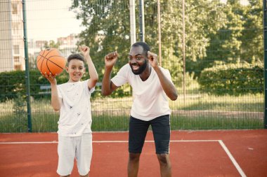 Genç babası ve oğlu parkın yakınındaki basketbol sahasında basketbol oynuyorlar. Beyaz tişört giyen adam ve çocuk. İkisinin de kolları havada, tezahürat yapıyorlar.