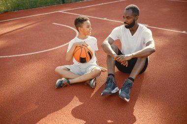 Genç baba ve oğlu parkın yakınındaki basketbol sahasında oturuyorlar. Beyaz tişört giyen adam ve çocuk. Konuşuyorlar ve ellerinde turuncu bir top tutuyorlar..