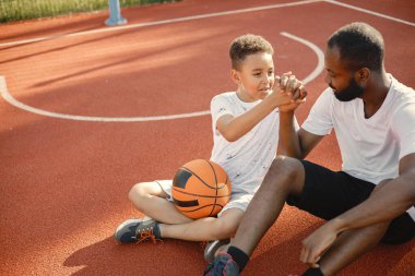 Genç baba ve oğlu parkın yakınındaki basketbol sahasında oturuyorlar. Beyaz tişört giyen adam ve çocuk. Konuşuyorlar ve el ele tutuşuyorlar..