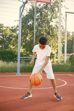 Genç çocuk parkın yanındaki basketbol sahasında duruyor. Beyaz tişört giyen bir çocuk. Çocuk basketbol topuyla oynuyor..