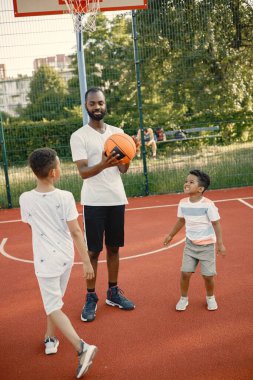 İki çok yönlü kardeş ve babaları parkın yakınındaki bir sahada basketbol oynuyorlar. Beyaz tişört giyen çocuklar. Ağabey, küçüğe basketbol oynamayı öğret..