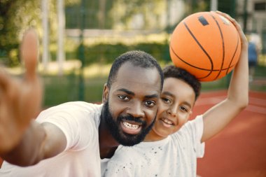 Genç baba ve oğlu parkın yanındaki basketbol sahasında duruyorlar. Beyaz tişört giyen adam ve çocuk. Baba oğul selfie çekiyorlar..