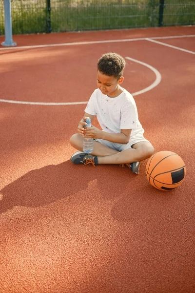 小男孩坐在公园附近的篮球场上 穿着白色T恤的男孩 男孩拿着一瓶水 — 图库照片