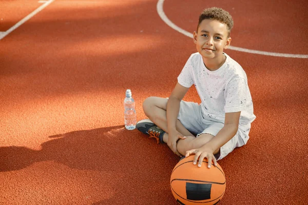 小男孩坐在公园附近的篮球场上 穿着白色T恤的男孩 男孩拿着一瓶水 看着镜头 — 图库照片