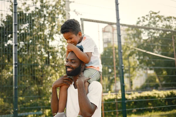 stock image Black father holding on shoulders his little son to help him to score a basket. They standing on a basketball court. Man and boy wearing white t-shirts.