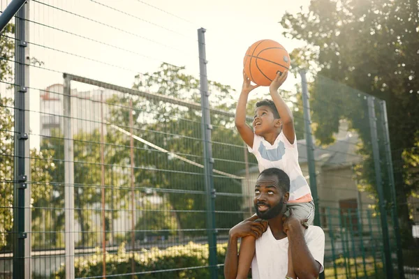 黑人父亲抱着他的小儿子帮他打篮球 他们站在篮球场上 穿着白色T恤衫的男人和男孩 — 图库照片